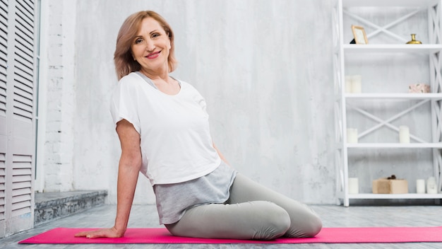 Donna senior adatta sorridente che si siede sulla stuoia di yoga che esamina macchina fotografica