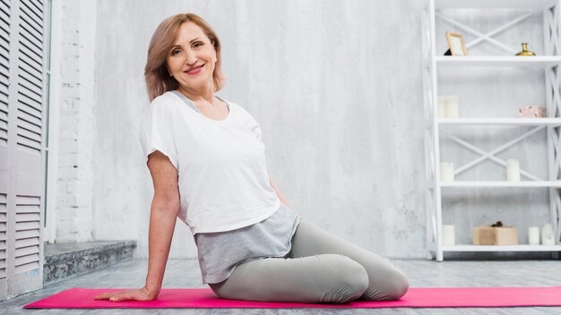 Smiling fit senior woman sitting on yoga mat looking at camera