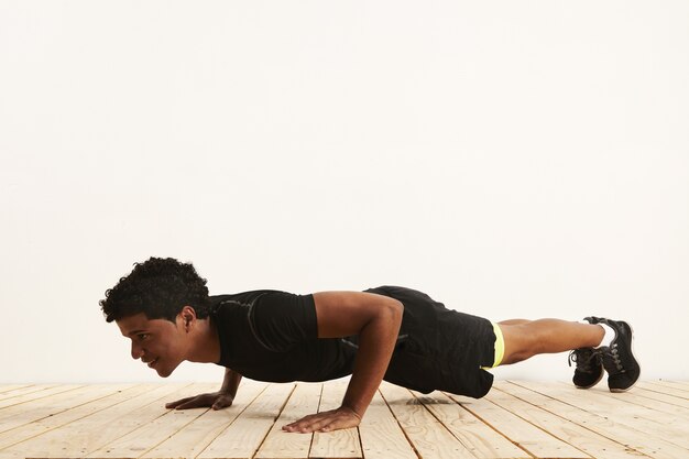 Free photo smiling fit young black athlete doing pushups on a light wooden floor against a white wall