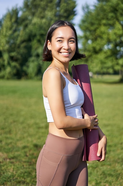 Free photo smiling fitness girl with rubber mat stands in park wearing uniform for workout and sport activities