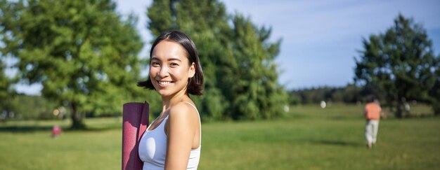 Free Photo smiling fitness girl with rubber mat stands in park wearing uniform for workout and sport activities