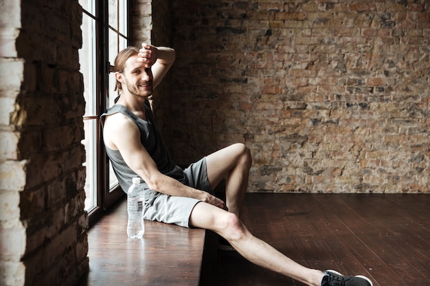 Free photo smiling fitness man resting at the windowsill with water bottle