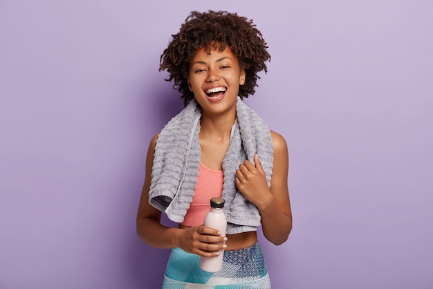Улыбающаяся фитнес-женщина в топе и леггинсах делает перерыв после тренировки, держит бутылку воды, вытирает пот полотенцем