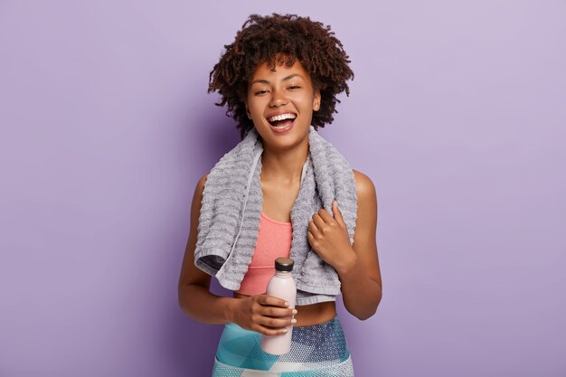 Smiling fitness woman in top and leggings takes break after training, holds bottle of water, wipes sweat with towel