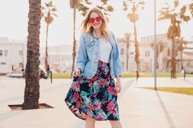 smiling flirting woman walking in city street in stylish printed skirt and denim oversize jacket wearing pink sunglasses