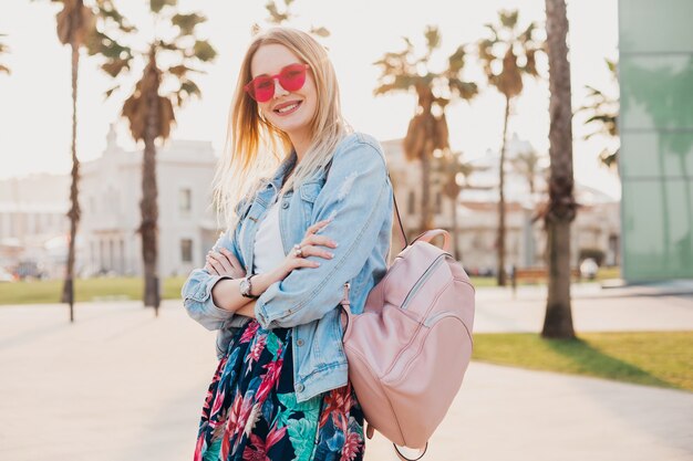 smiling flirting woman walking in city street in stylish printed skirt and denim oversize jacket wearing pink sunglasses