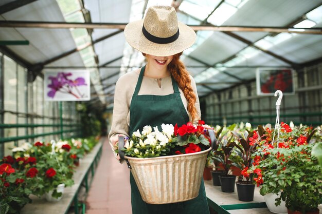 Улыбающийся флорист в фартуке стоит и держит цветы в металлической корзине, закрывая лицо шляпой в теплице