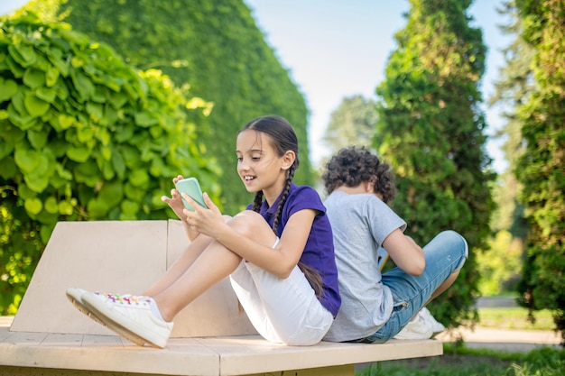 Улыбающаяся сосредоточенная девушка сидит на скамейке рядом с кудрявым мальчиком и пользуется своим мобильным телефоном