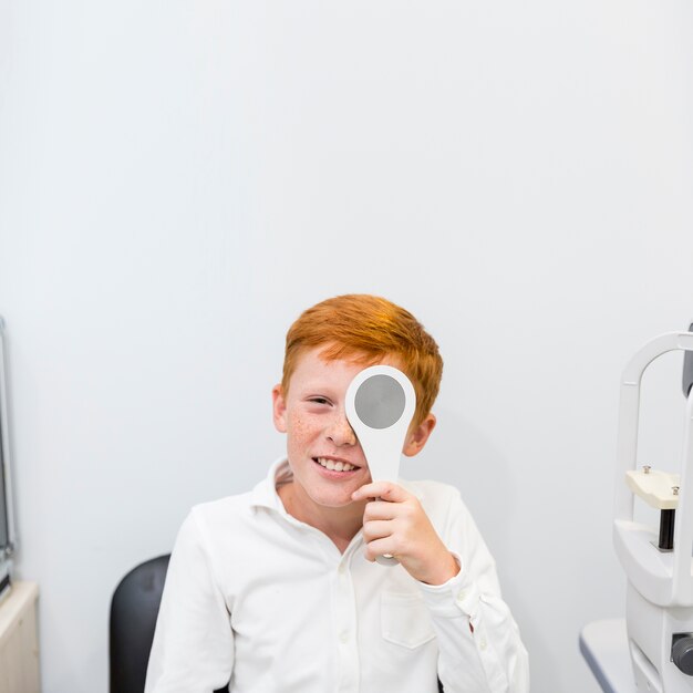 Smiling freckle boy holding occluder in front of his eye in ophthalmological clinic