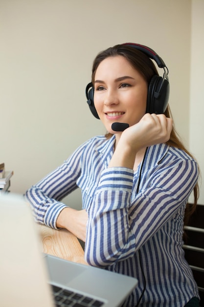 Smiling freelance operator working online with headsets and a laptop in a desktop at office. Cheerful call centre girl working from home talking with customer. Vertical