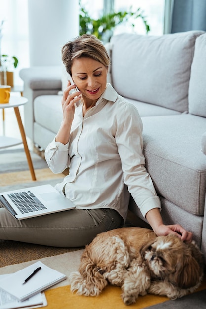 Smiling freelance worker with a dog communicating on cell phone while working on laptop at home