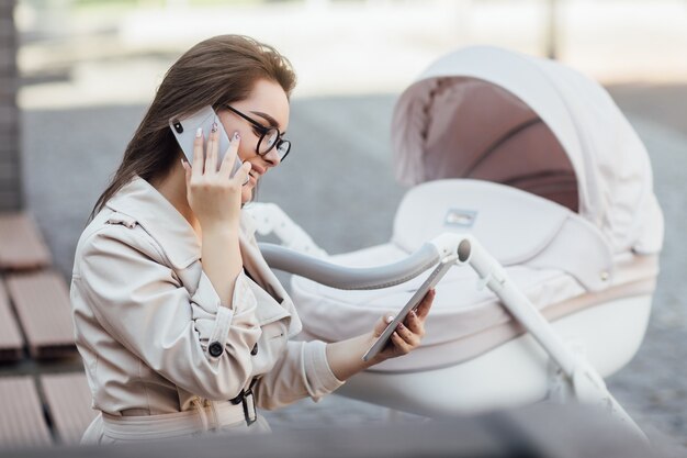 Smiling freelancer mom working with phone on bench near baby stroller in park.