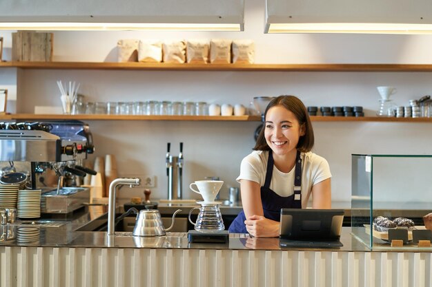 カウンター醸造フィルター コーヒー管理で働くエプロンを身に着けている笑顔のフレンドリーなアジアの女の子バリスタ