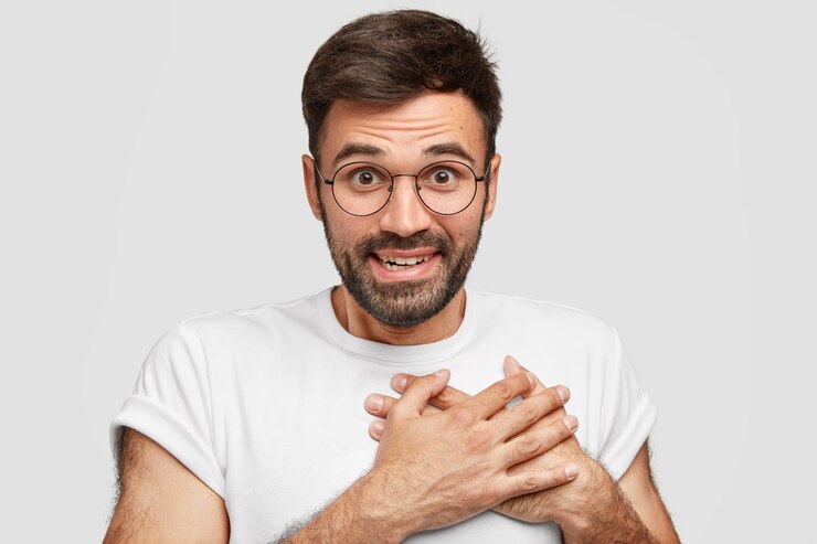 Free Photo smiling friendly looking male with stubble, keeps hands on chest, expresses gratitude, has happy expression, wears casual t-shirt, has dark beard, isolated on white wall. acknowledgement concept