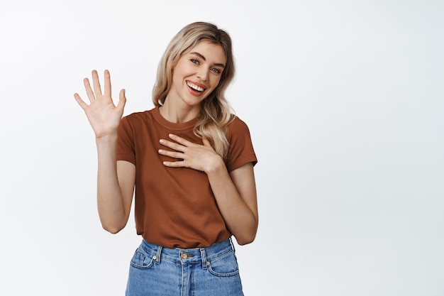 Smiling friendly woman raising hand and hold arm on heart introduce herself saying hello standing in tshirt against white background