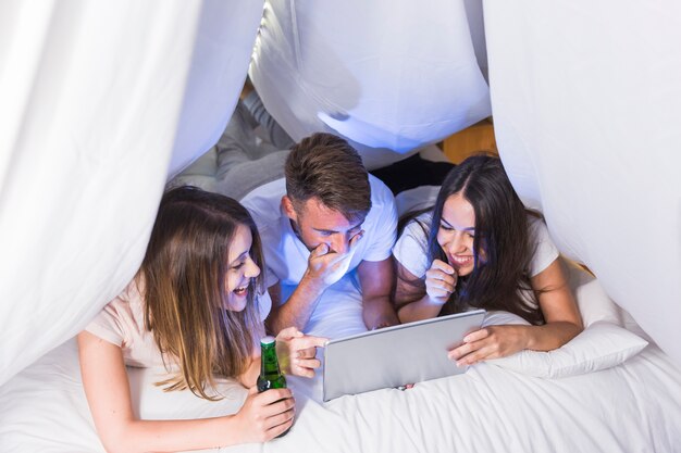 Smiling friends lying on bed looking at digital tablet