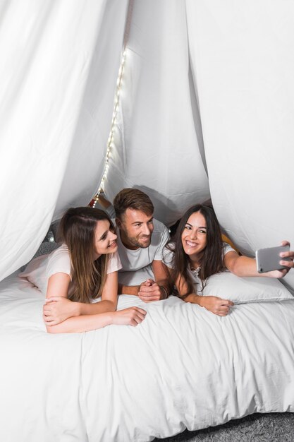 Smiling friends lying on bed taking selfie from cellphone