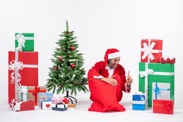 Smiling funny excited young man dressed as Santa claus with gifts and decorated Christmas tree sitting on the ground pointing above on white background