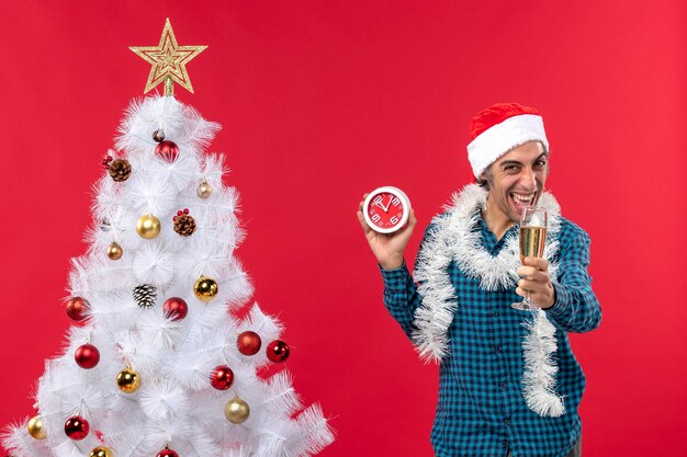 Smiling funny young guy with santa claus hat and raising a glass of wine and holding clock standing near Xmas tree on red