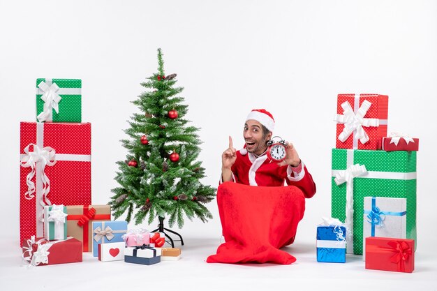 笑顔の面白い若い男は、地面に座って、贈り物や飾られたクリスマスツリーの近くの上を指している時計を保持して新年やクリスマス休暇を祝います
