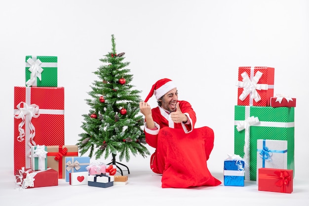 Smiling funny young man dressed as Santa claus with gifts and decorated Christmas tree sitting on the ground making ok gesture on white background