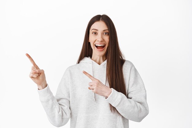 Smiling genz girl look fascinated pointing fingers at upper left corner showing logo or website link demonstrate banner recommending product standing happy against white background