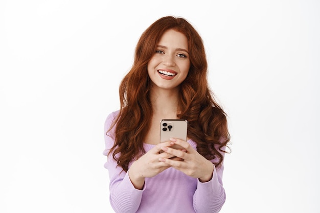 Smiling ginger girl chatting, holding smartphone as if shopping online, looking happy while messaging, standing in blouse against white background