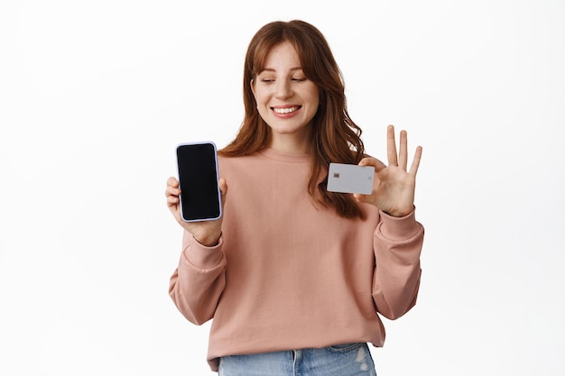 Smiling ginger girl showing mobile phone screen and credit card, standing against white background.
