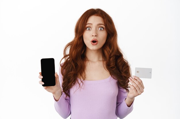 Smiling ginger woman holding smartphone and credit card