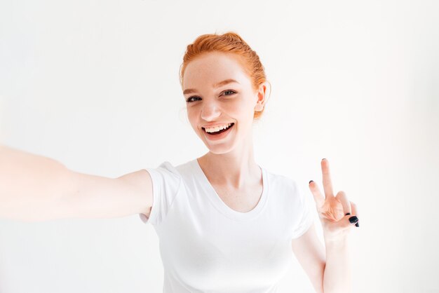 Smiling ginger woman in t-shirt making selfie and showing peace gesture