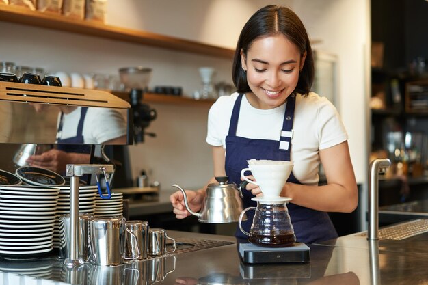 笑みを浮かべて少女バリスタ アジア バーテンダーの後ろのカフェでやかん醸造フィルター コーヒーから水を注ぐ