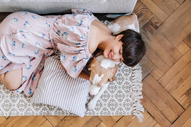 美しいロングドレスの笑顔の女の子は目を閉じてビーグル犬に横たわっています。彼女のペットと一緒にカーペットの上で冷やしている魅力的な笑っている女性の上からの肖像画