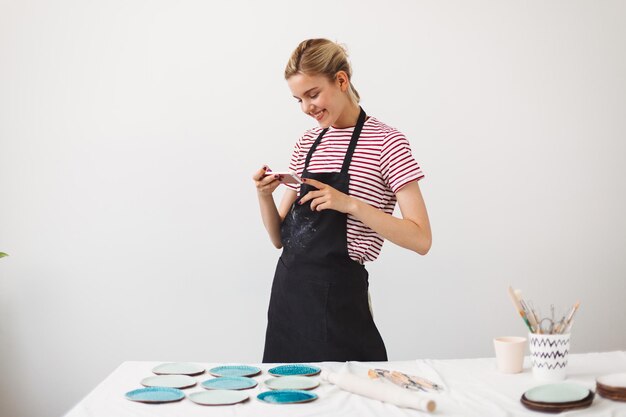 Smiling girl in black apron and striped T-shirt happily taking photos of handmade plates on her cellphone at pottery studio