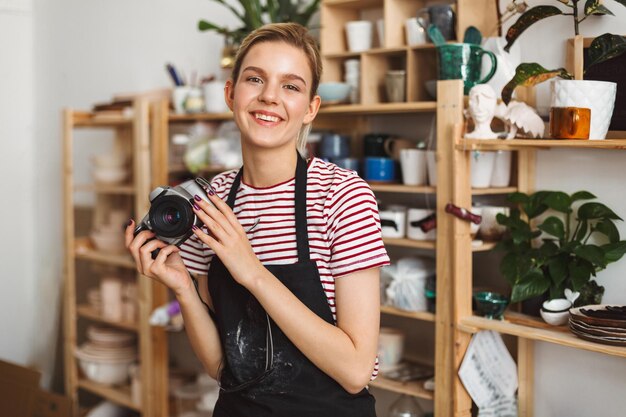 Free photo smiling girl in black apron and striped t-shirt holding camera in hands happily looking in camera at pottery studio