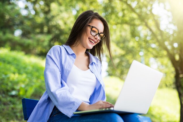 Улыбающаяся девушка в синей футболке сидит на скамейке в парке и использует свой ноутбук