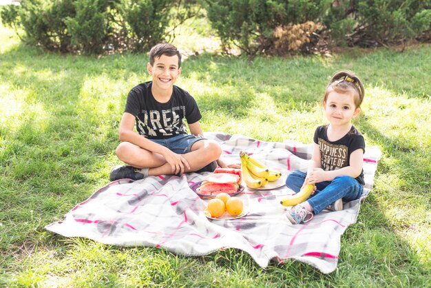 smiling girl and boy sitting on blanket over the green grass with fruits