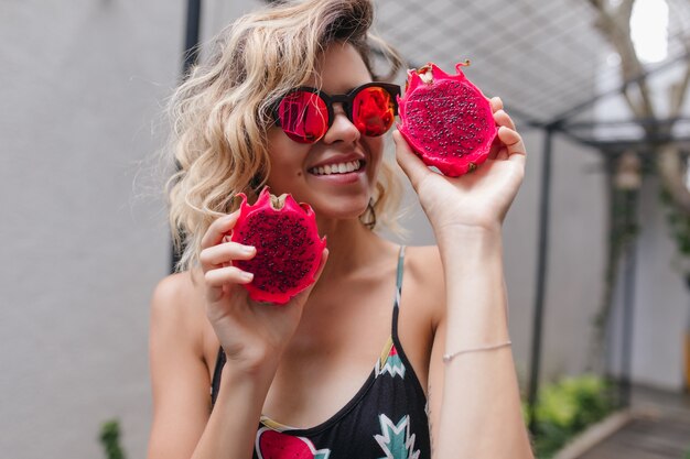 Smiling girl in bracelet posing with dragon fruit. Photo of lovely curly woman in sunglasses holding red pitaya.