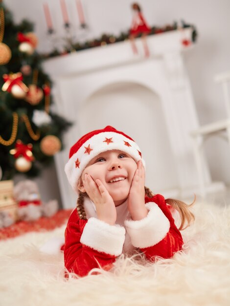 Smiling girl dressed in santa claus lying on the floor with hands on face