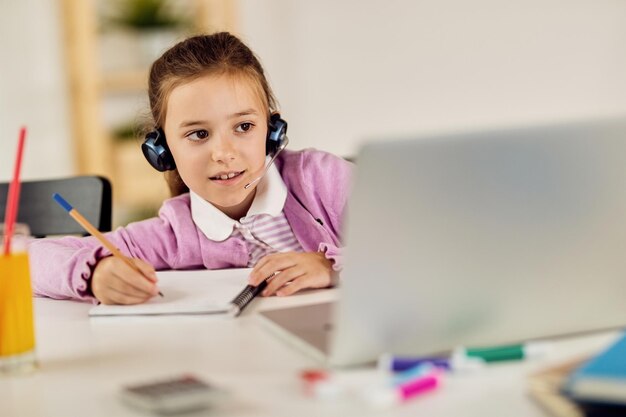 Smiling girl elearning and writing in notebook while homeschooling