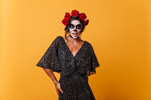 Smiling girl emotionally posing in Mexican skeleton mask. Model with roses in her hair laughs on orange wall.