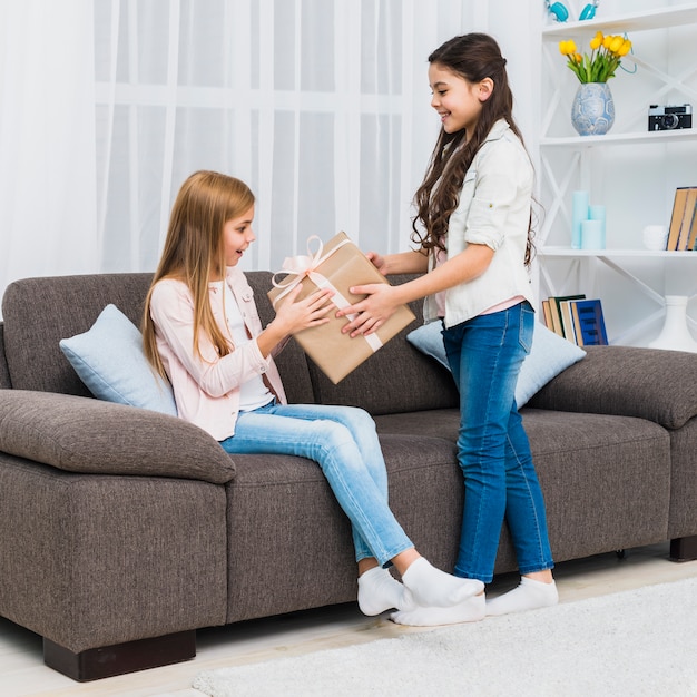 Smiling girl giving present to her friend sitting on sofa at home