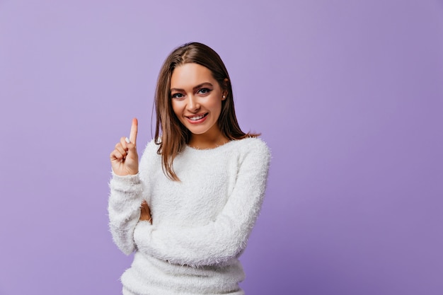 Smiling girl of good appearance remembered happy memory and raises her index finger. Portrait of cute european model in soft sweater