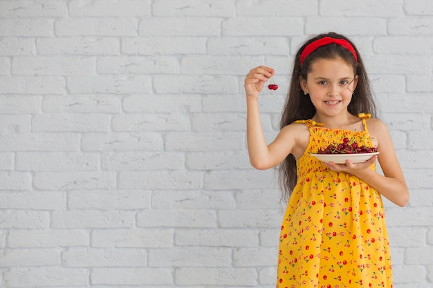Smiling girl holding cherries in plate standing against brick wall