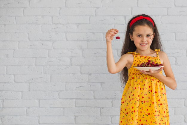 Smiling girl holding cherries in plate standing against brick wall