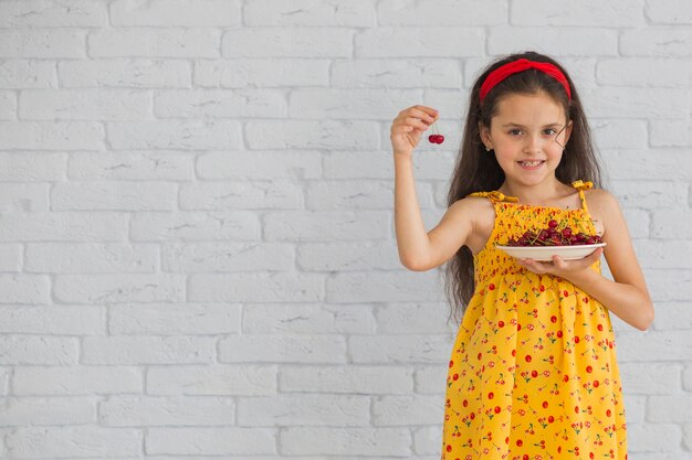 Улыбающаяся девочка, держащая вишни в тарелке, стоящей против кирпичной стены