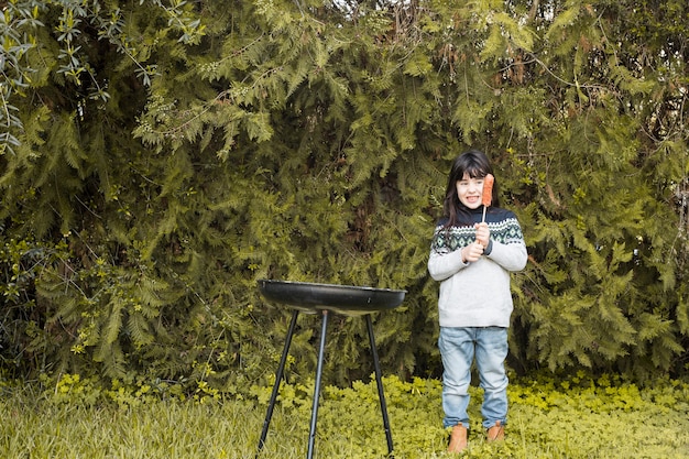 携帯バーベキューの近くに焼きたてのソーセージを握っている笑顔の女の子