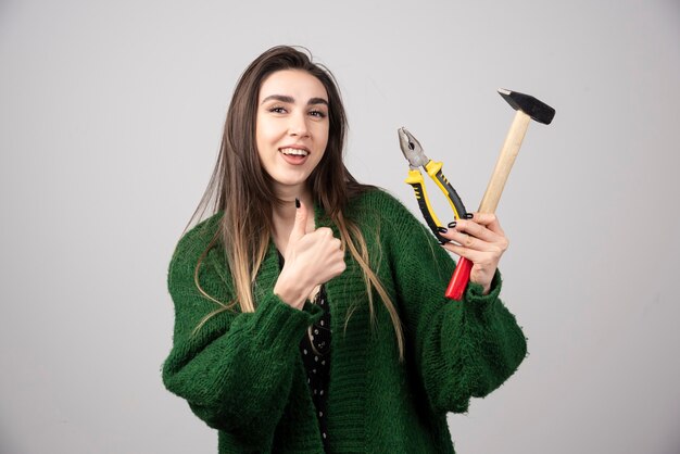 Smiling girl holding a hammer with pliers and showing a thumb up.