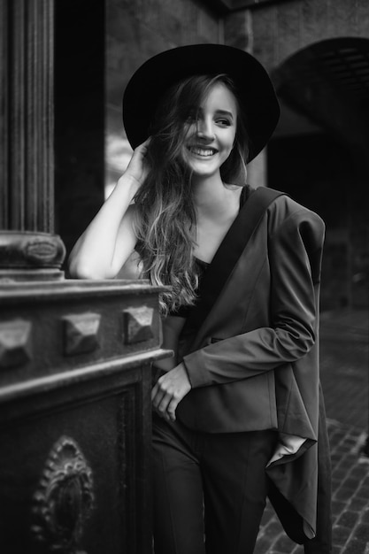 Smiling girl holding a hat standing in the street. Fashion model in the city. Black and white fashion phography. Stylish woman wearing siut and hat.