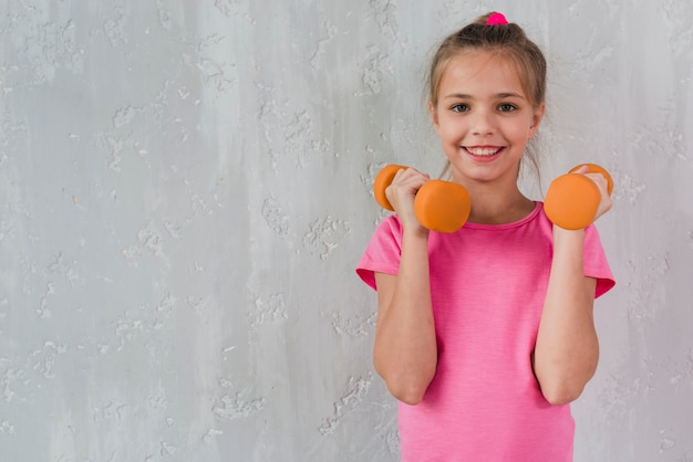 Smiling girl holding orange dumbbell in front of concrete wall