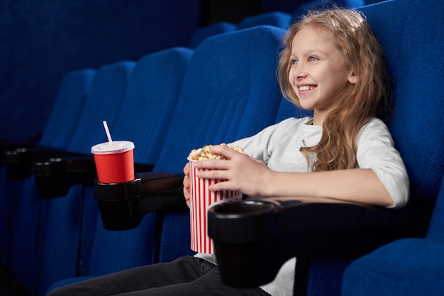 Smiling girl holding popcorn bucket, sitting in cinema.